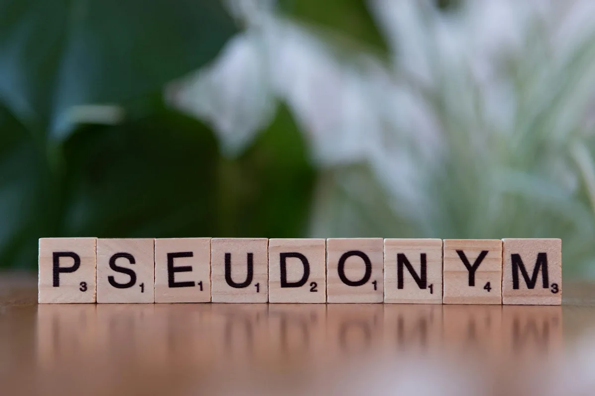 Scrabble tiles spelling "PSEUDONYM" with blurred green and white foliage in background