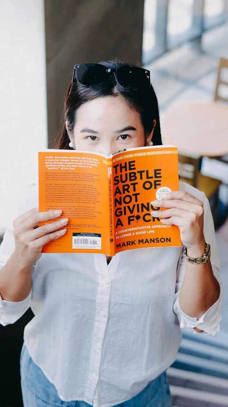 Woman in white shirt holding open "The Subtle Art of Not Giving a F*ck" book by Mark Manson, covering her face