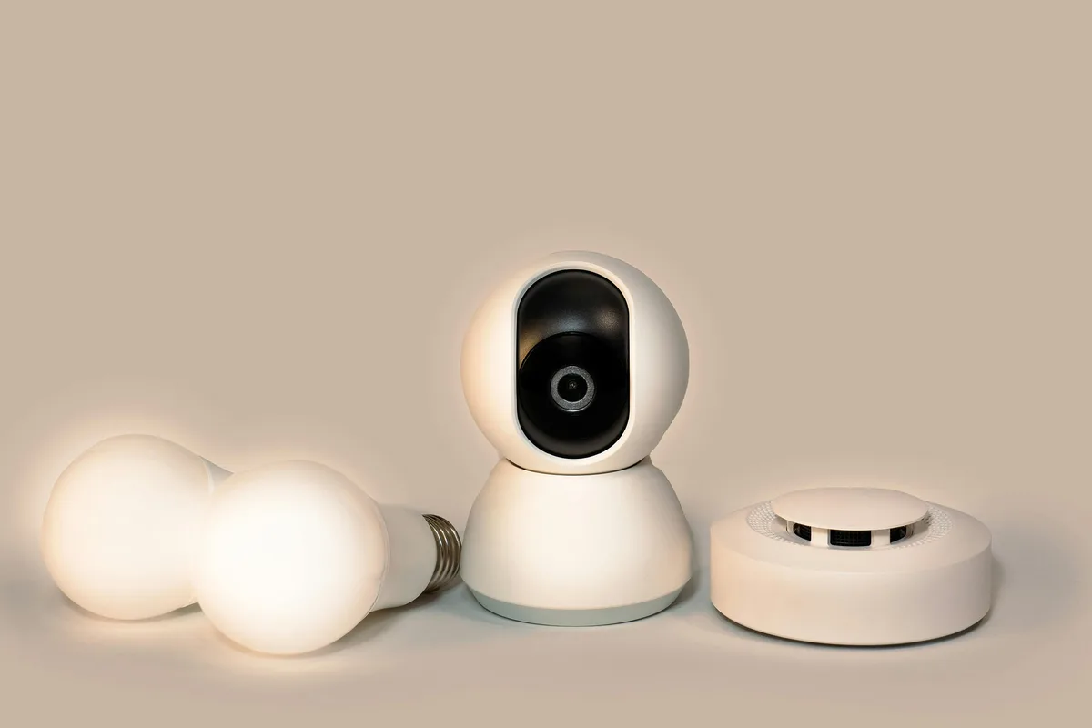 Smart home devices including white LED bulbs, a pan-tilt security camera, and a smoke detector on neutral background