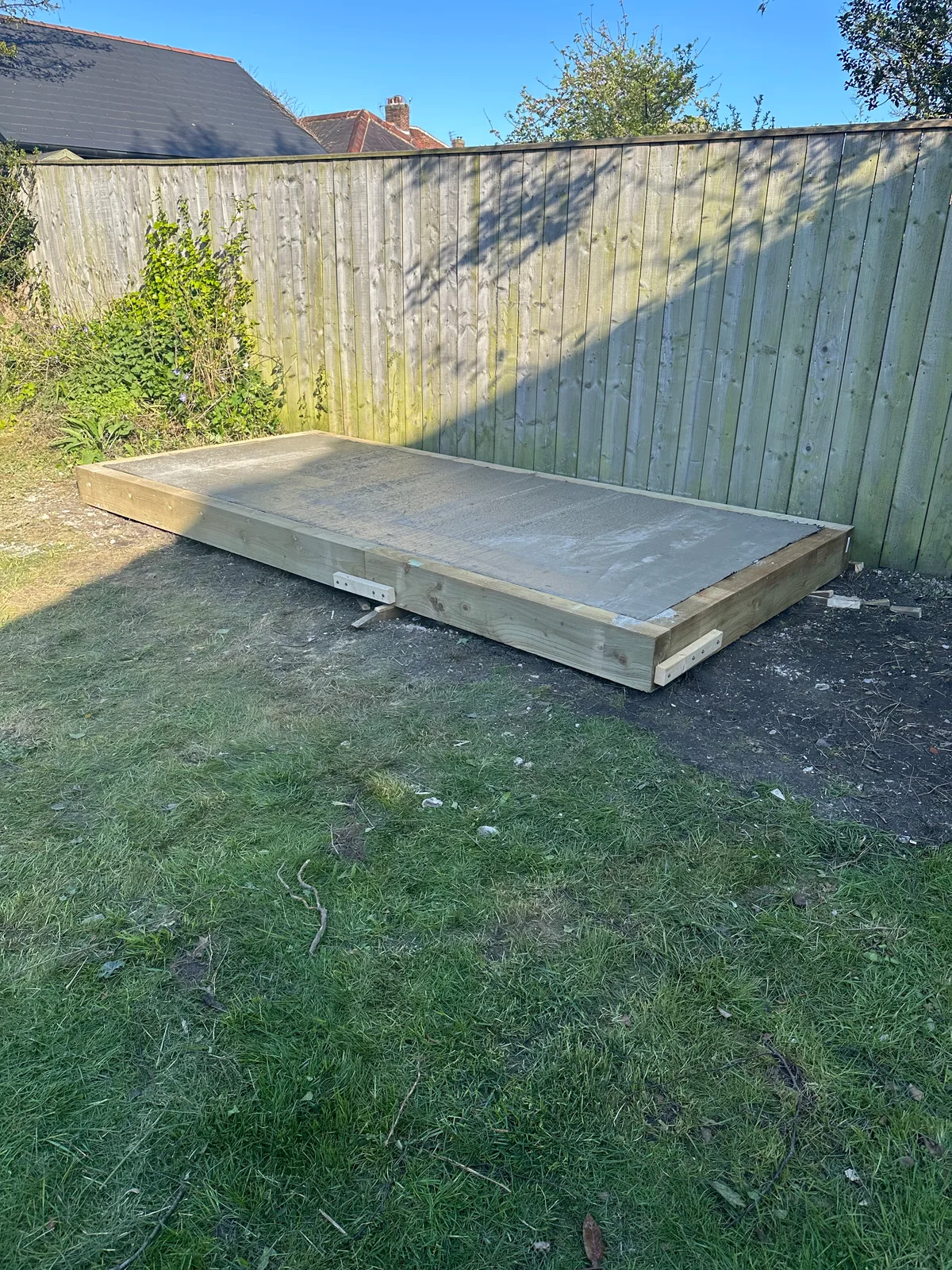 A concrete shed base framed in timber, sitting on the lawn in front of a garden fence