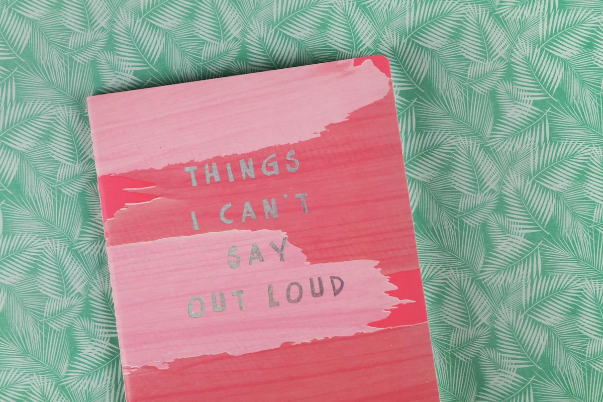 A pink notebook with gray text reading "Things I Can't Say Out Loud" on a green leaf-patterned background.