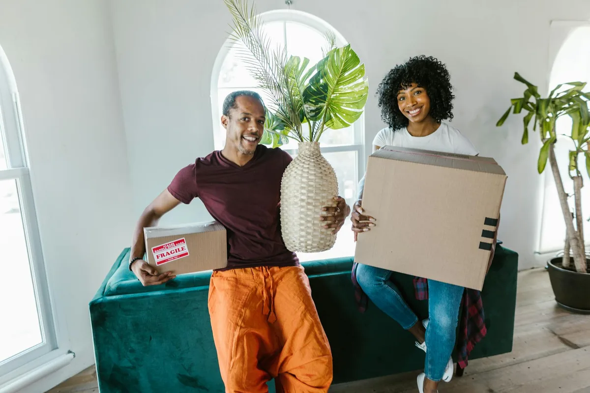 Happy couple holding moving boxes in bright apartment with potted plants