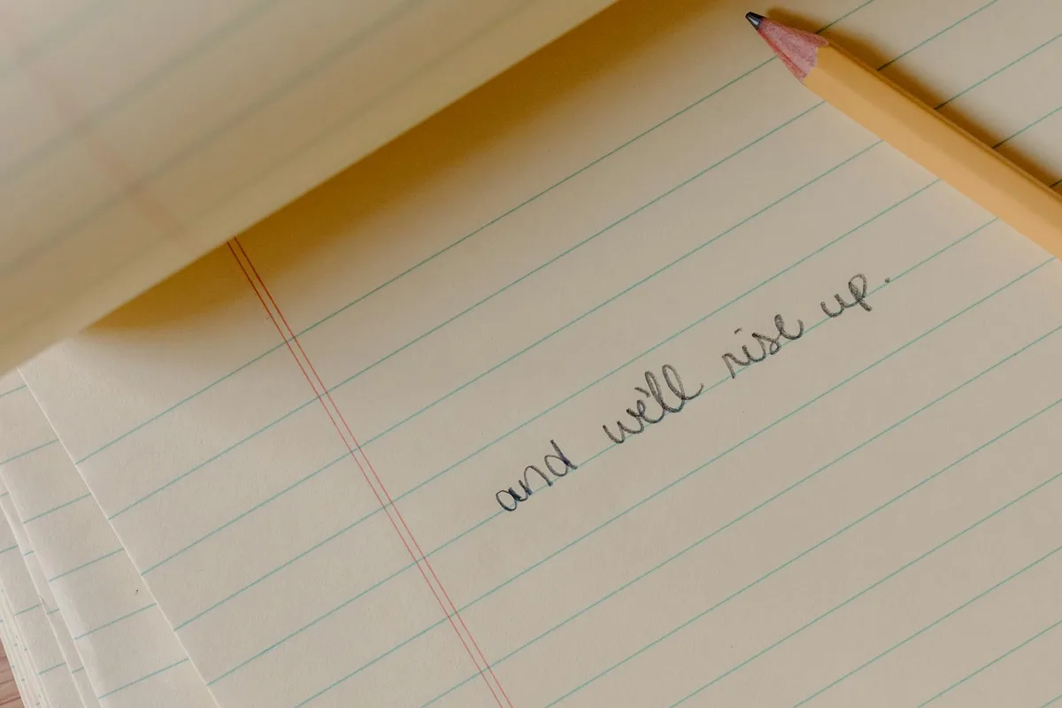 A sharpened pencil rests on lined notebook paper with handwritten text reading "and well rise up"