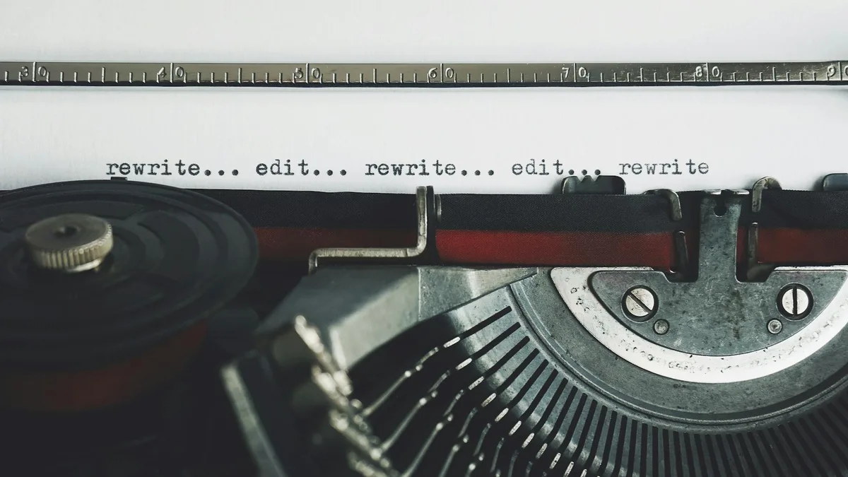 Typewriter with paper showing repeated text "rewrite... edit... rewrite... edit... rewrite" against a ruler backdrop